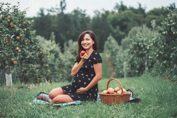 Happy healthy pregnancy. Portrait of pregnant young brunette Caucasian woman sitting on grass eating apple. Beautiful expecting mom lady on farm at countryside, rustic style.