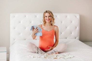 Çocuk giyim romper tutan gülümseyerek, beyaz beyaz sarışın hamile kadın portre. Mutlu genç orta yaş kadın kadın yatakta yatak odasında oturmuş bekliyor. Yeni doğan bebek hazırlıkları.