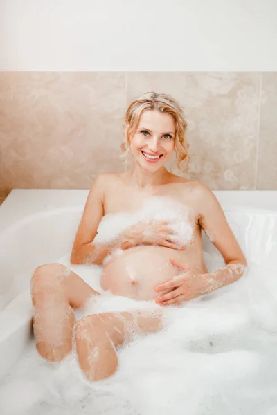 Happy healthy pregnancy. Portrait of pregnant blonde Caucasian woman washing in bathroom. Beautiful expecting mom lady taking morning bath with foam and having fun.