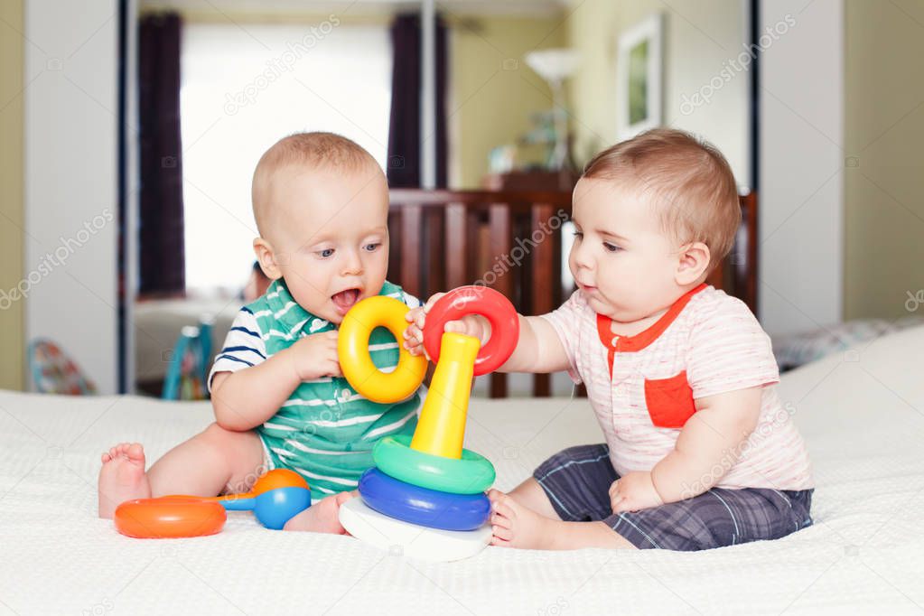 Group portrait of two white Caucasian cute funny baby boys sitting together on bed playing stacking rings toy. Friendship childhood concept. Best friends forever. Team work building