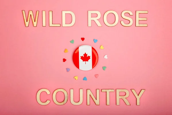 Happy Canada Day greeting card with wooden letters. Alberta provincial license slogan saying wild rose country and Canadian flag with many hearts on living coral pink background.