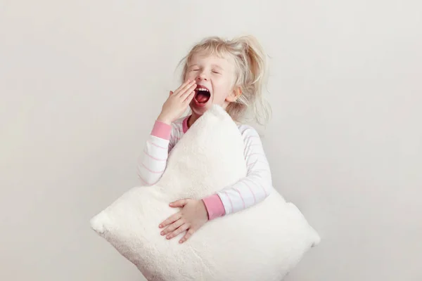 Healthy sleep concept. Cute blonde Caucasian girl child in pink pajamas with closed eyes yawning covering wide open mouth with palm. Sleepy kid with messy hair holding white soft pillow