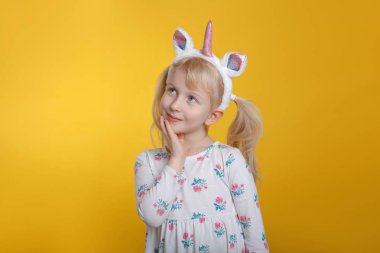 Sevimli sevimli dalgın Beyaz sarışın kız beyaz elbise giyen tek boynuzlu at kafa bandı boynuz ve kulakları hayal düşünme. Komik çocuk çocuk duygu ayakta Studio Sarı zemin üzerine ifade