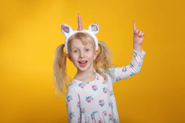 Sevimli sevimli gülümseyen Beyaz sarışın kız beyaz elbise giyen tek boynuzlu at kafa bandı boynuz ve kulakları Studio Sarı zemin üzerine. Duygu ifade ve gösteren işaret komik çocuk çocuk