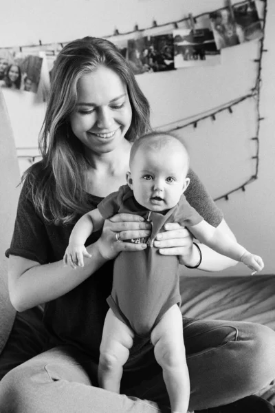Black-and-white portrait of Caucasian woman mother holding baby boy girl child sitting on bed at home. Authentic candid family lifestyle. Happy motherhood and parenting. Film grain noise added