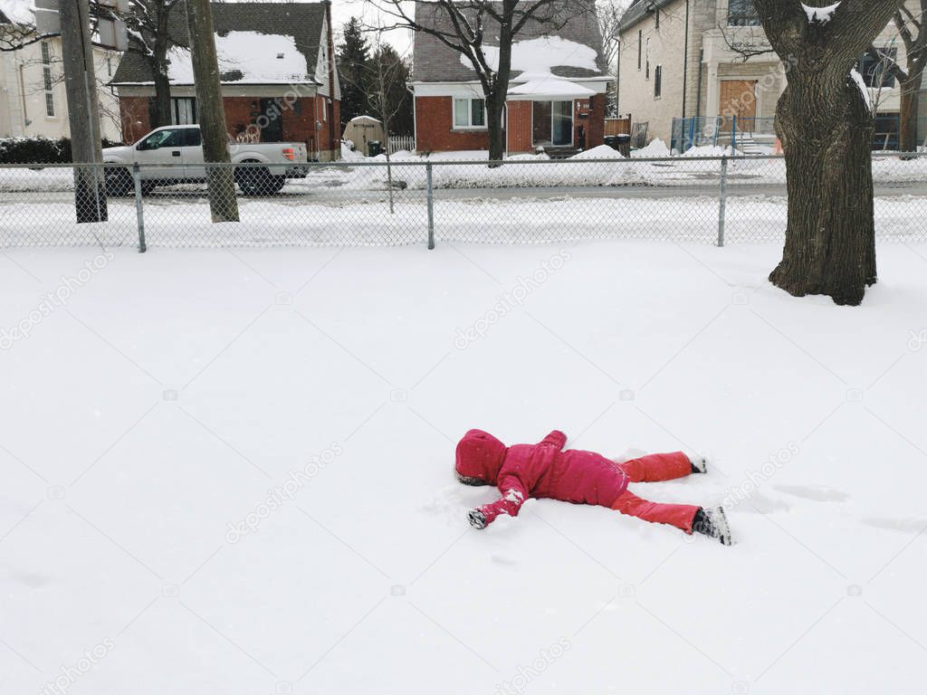Funny preschool child girl in warm clothes pink red jacket lying in snow during cold winter day with her face down. Upset kid doesn't want to go to school. Naughty cranky kid playing outdoors.