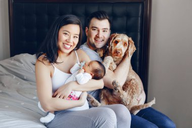 Gülen Çinli Asyalı anne ve melez baba ile karışık ırk yeni doğan bebek oğlu kızı ve büyük evcil hayvan köpek. Yatak odasında mutlu bir aile. Ev yaşam tarzı otantik doğal an. 