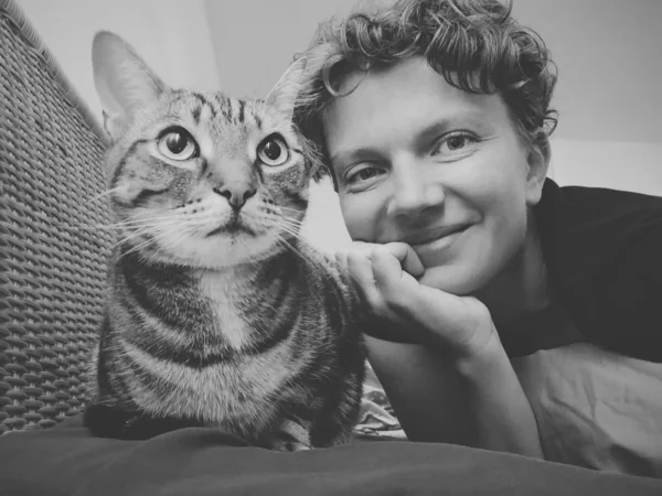 Black and white selfie portrait of cat and its owner girl lying on couch sofa looking in camera. Smiling Caucasian woman with kitten pet feline animal. Authentic style real person lifestyle.