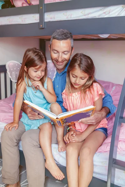 Family of three members people sitting on bed in bedroom reading book. Father and daughters girls at home spending time together. Parent talking communicate to children. Real people lifestyle