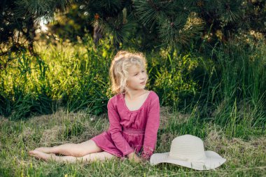 Gün batımında park açık zemin çim üzerinde oturan dağınık dağınık saçları ile kırmızı pembe elbiseli sevimli güzel sarışın Kafkas kız Portresi. Mutlu sevimli yalınayak çocuk yaz zevk. 