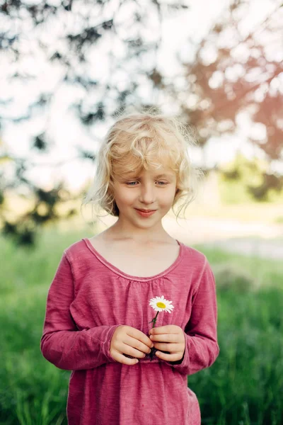 Portrait of cute beautiful blonde Caucasian girl in red pink dress holding white daisy flower in her hands. Happy adorable child kid with messy untidy hair enjoying summertime. Lifestyle childhood.