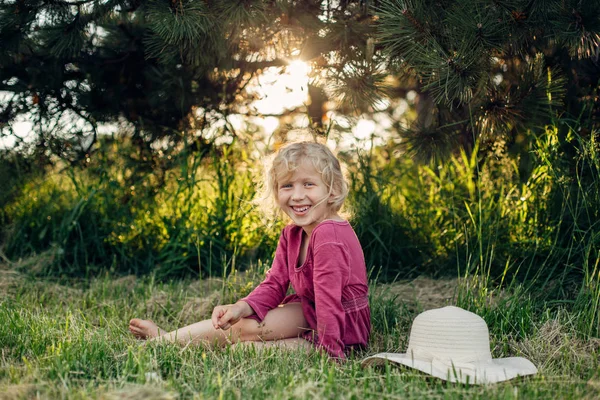 Gün batımında park açık zemin çim üzerinde oturan dağınık dağınık saçları ile kırmızı pembe elbiseli sevimli güzel sarışın Kafkas kız Portresi. Mutlu sevimli yalınayak çocuk yaz zevk. 