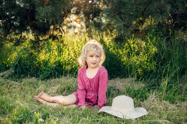 Gün batımında park açık zemin çim üzerinde oturan dağınık dağınık saçları ile kırmızı pembe elbiseli sevimli güzel sarışın Kafkas kız Portresi. Mutlu sevimli yalınayak çocuk yaz zevk. 