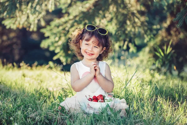 可爱的可爱的白种幼儿女婴坐在草地上 吃浆果水果 有趣的孩子与太阳镜在公园有健康的小吃餐 夏季美味的手指食品为孩子们 — 图库照片