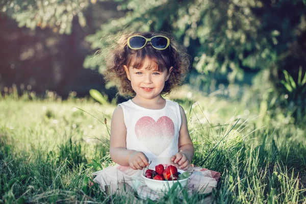 かわいい愛らしい白人の幼児の女の子は草の上に座って ベリーの果物を食べています 健康的なスナックの食事を持つ公園でサングラスを持つ面白い子供 子供のための夏のおいしいおいしい指の食べ物 — ストック写真