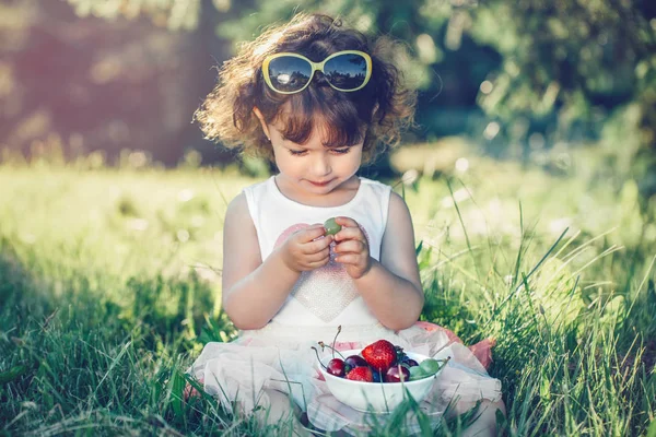 かわいい愛らしい白人の幼児の女の子は草の上に座って ベリーの果物を食べています 健康的なスナックの食事を持つ公園でサングラスを持つ面白い子供 子供のための夏のおいしいおいしい指の食べ物 — ストック写真
