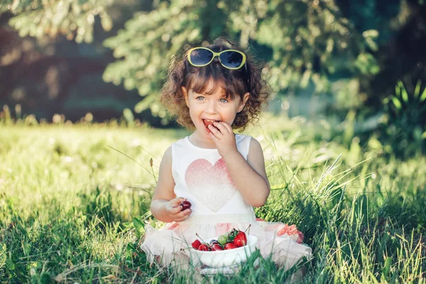 かわいい愛らしい白人の幼児の女の子は草の上に座って ベリーの果物を食べています 健康的なスナックの食事を持つ公園でサングラスを持つ面白い子供 子供のための夏のおいしいおいしい指の食べ物 — ストック写真