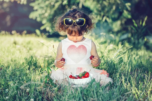 可爱的可爱的白种幼儿女婴坐在草地上 吃浆果水果 有趣的孩子与太阳镜在公园有健康的小吃餐 夏季美味的手指食品为孩子们 — 图库照片