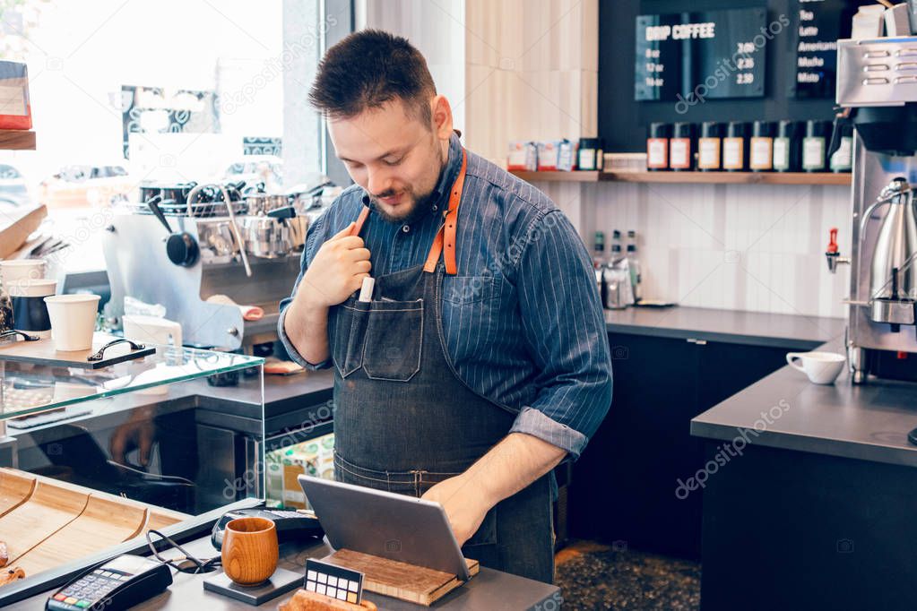 Young bearded Caucasian male man cashier barista in restaurant cafe at work. Seller using touch pad for accepting client customer payment purchase. Small business of coffee shop cafeteria. 