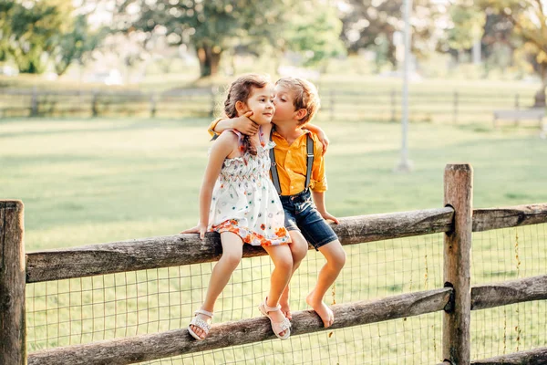 两个可爱的有趣的白人儿童男孩女孩坐在外面的公园围栏 朋友兄弟姐妹的孩子拥抱亲吻在夏天的日落 浪漫爱情和友谊自然生活方式真实时刻 — 图库照片