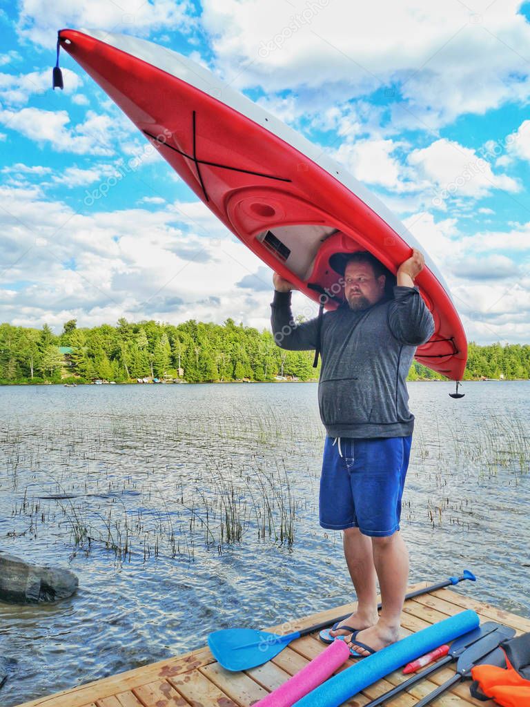 Fat stout Caucasian man standing on lake river wooden dock and holding kayak boat on his head at sunny summer day. Funny concept of physical activity and sports for adults to loose weight.