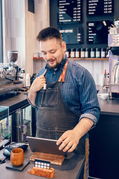 Young bearded Caucasian male man cashier barista in restaurant cafe at work. Seller using touch pad for accepting client customer payment purchase. Small business of coffee shop cafeteria.