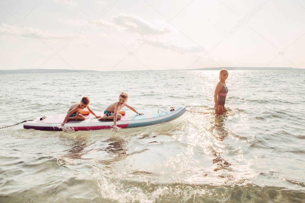 Caucasian woman parent riding kids children boys on paddle sup surfboard in water. Modern outdoor summer fun family activity. Individual aquatic recreation sport hobby. Healthy lifestyle.