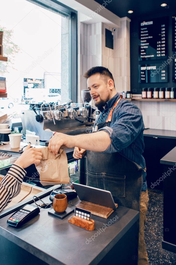 Smiling bearded Caucasian man cashier barista in restaurant cafe at work. Seller giving client customer her purchase food in brown organic paper package bag. Small business of coffee shop cafeteria. 