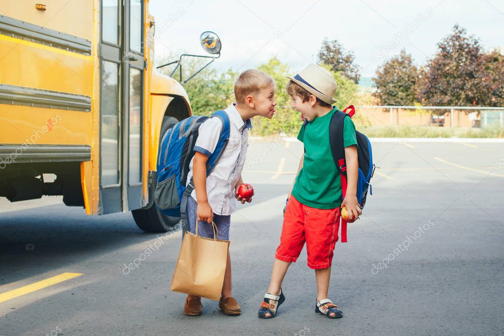 Two funny happy Caucasian boys students kids showing tongues each other near yellow bus on 1 September day. Education and back to school concept. Children pupils ready to learn and study. 