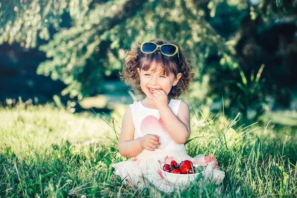 かわいい愛らしい白人の幼児の女の子は草の上に座って ベリーの果物を食べています 健康的なスナックの食事を持つ公園でサングラスを持つ面白い子供 子供のための夏のおいしいおいしい指の食べ物 — ストック写真