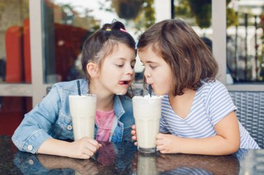 Two funny Caucasian little preschool sisters siblings drink milk shakes in cafe. Friends girls having fun together. Cold summer desserts for kids. Happy authentic childhood lifestyle.  clipart