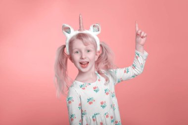 Beyaz elbiseli, tek boynuzlu at boynuzuyla pembe mercan arka planda gülümseyen, sevimli, sevimli, beyaz, sarışın kız. Komik çocuk duygularını ifade ediyor ve işaret ediyor..