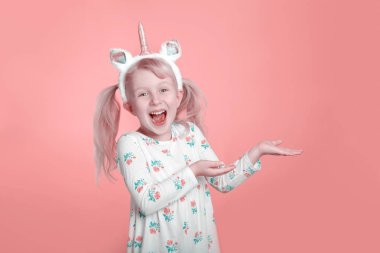 Beyaz elbiseli, sevimli, sevimli, gülümseyen, beyaz bir kız. Stüdyoda tek boynuzlu at boynuzu takıyor. Mercan pembesi arka planda. Komik çocuk duygularını ifade ediyor ve işaret ediyor..
