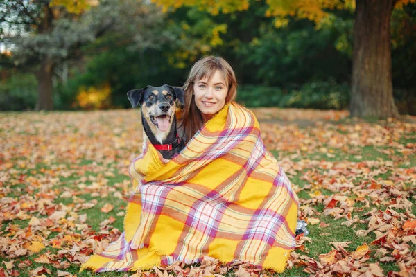 Woman wrapped in woollen blanket with dog in autumn fall park. Beautiful happy young Caucasian woman sitting on ground hugging domestic canine animal pet. Best friends forever.