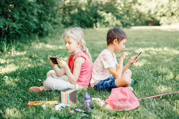 Erkek ve kız dışarıda akıllı telefonlarda oyun oynuyorlar. Çocukların dijital cihaz ekran bağımlılığı. Çocuk arkadaşlar parkta çimlerin üzerinde oynuyorlar. Çağdaş yalnızlık sorunu..