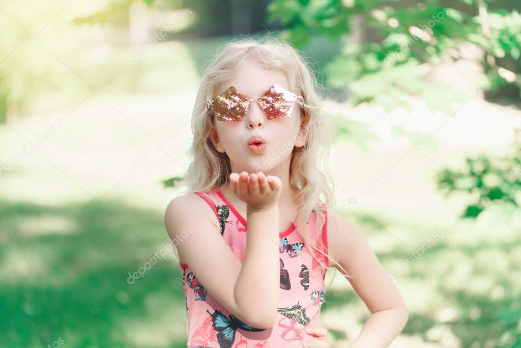 Young girl posing in fancy pink pentagonal shaped sunglasses outdoor. Cute adorable stylish Caucasian child with long blonde hair sending air kiss. Cool hipster kid having fun. 