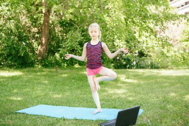 İnternette yoga yapan beyaz bir kız çocuğu. İnternetteki video spor antrenmanı. Çocuk dizüstü bilgisayarla spor yapmayı öğreniyor. Yeni normal. Coronavirus 'ta sosyal uzaklık. Stres atma kavramı.