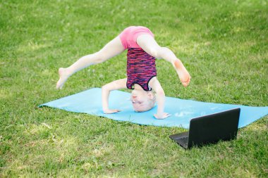Kız çocuğu dışarıda spor yapıyor. İnternette. İnternette parkta gerinen video yogası. Çocuk arka bahçede dizüstü bilgisayarla jimnastik öğreniyor. Yeni normal. Coronavirus 'ta sosyal uzaklık.