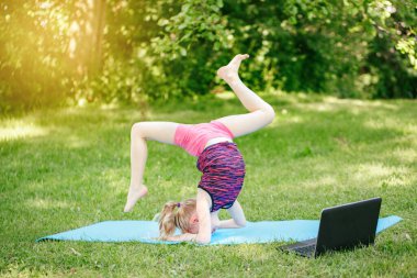 Kız çocuğu dışarıda spor yapıyor. İnternette. İnternette parkta gerinen video yogası. Çocuk arka bahçede dizüstü bilgisayarla jimnastik öğreniyor. Yeni normal. Coronavirus 'ta sosyal uzaklık.