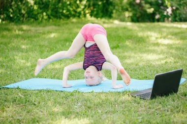 Kız çocuğu dışarıda spor yapıyor. İnternette. İnternette parkta gerinen video yogası. Çocuk arka bahçede dizüstü bilgisayarla jimnastik öğreniyor. Yeni normal. Coronavirus 'ta sosyal uzaklık.