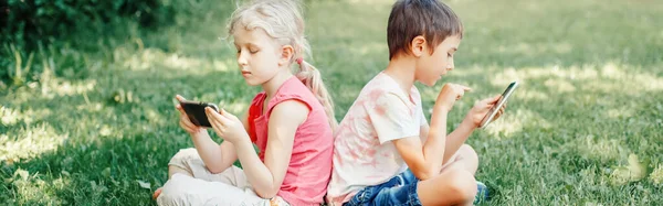 Children boy and girl friends play games on smartphones outdoor. Kids digital gadget screen addiction. Contemporary problem of loneliness together. Web banner header for website.