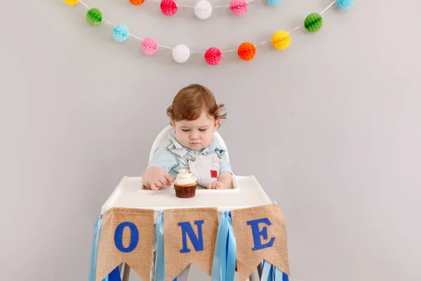 かわいい愛らしい悲しい動揺白人の赤ちゃん男の子自宅で彼の最初の誕生日を祝う 高椅子テーブルに座っている幼児の幼児はおいしいカップケーキデザートを食べています 幸せな誕生日のライフスタイルのコンセプト — ストック写真