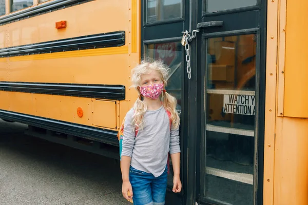 Sad girl student with face mask near locked yellow bus. Closed school during pandemic lockdown and quarantine shutdown. Education and back to school. New normal in coronavirus.