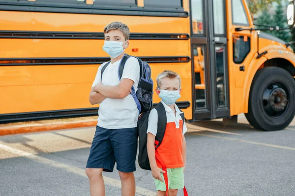 黄色いバスの近くにフェイスマスクをした悲しい兄弟学生 顔に個人用保護具付きの子供 教育と9月に学校に戻る コロナウイルスの新常態 — ストック写真