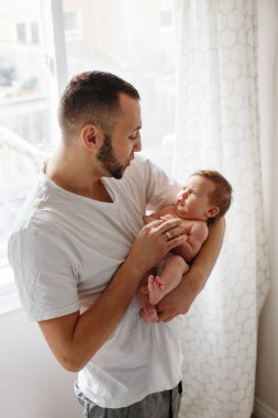 Yeni doğmuş bebeği tutan mutlu beyaz bir baba. Erkek ebeveyn sallanıyor ve kendi elleriyle çocuk kızıyla konuşuyor. Otantik yaşam tarzı samimi anı. Babalar Günü tatili.