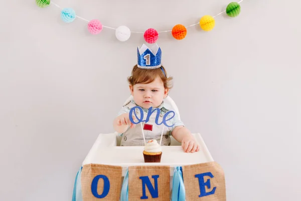 祝你生日快乐 可爱的白种人男婴身穿蓝色王冠在家里庆祝第一个生日 小孩子坐在高高的椅子上 吃着美味的纸杯蛋糕甜点 上面加了单词 Topper 主题活动 — 图库照片