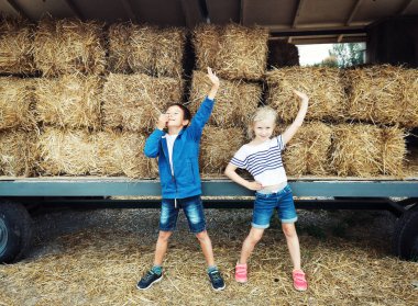 Çiftlikte saman yığınının önünde poz veren mutlu komik çocuklar. Sonbahar sonbaharı çiftlik parkı mevsimlik dekorasyonu. Kuru samanlı büyük bir araba. Mutlu çocukluk hayatı.