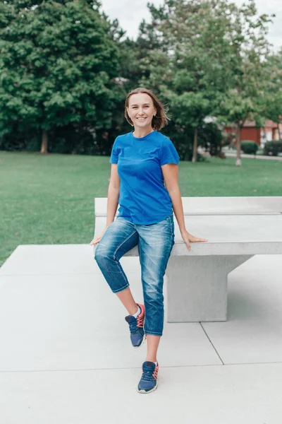 Beautiful happy smiling sporty middle age woman in blue t-shirt and jeans park. Caucasian woman with short hair bob in park outdoor. Fit and slim sporty female person after workout.