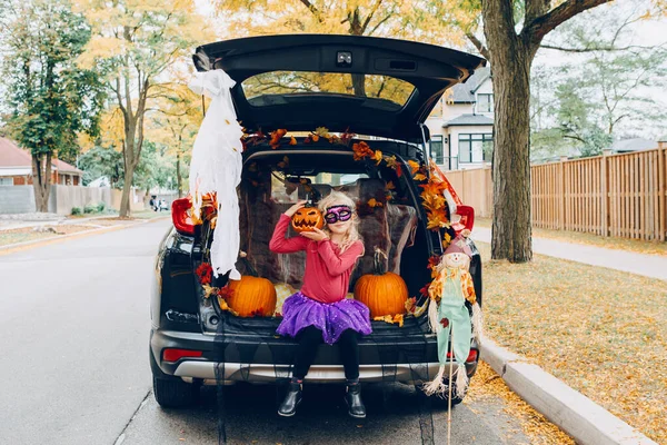 Trick or trunk. Child girl celebrating Halloween in trunk of car. Kid with red carved pumpkin celebrating traditional October holiday outdoor. Social distance and safe alternative celebration.