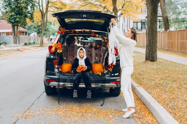 Trick or trunk. Child boy with red pumpkin for Halloween sitting in trunk of car. Mother decorating car for traditional October holiday outdoor. Social distance and safe alternative celebration.